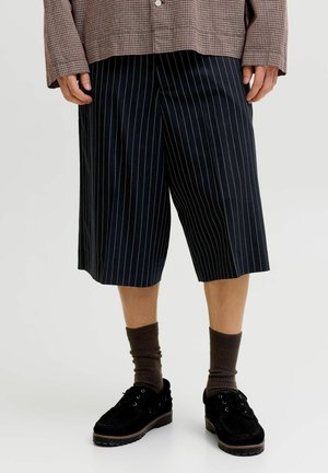 Personne portant un short large à fines rayures noires, des chaussettes marron, des chaussures à plateforme noires et une veste marron à carreaux, debout sur un fond uni.