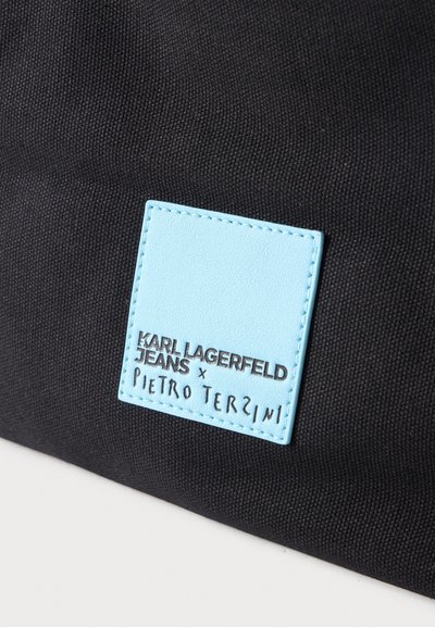 Близък план на черна текстурирана чанта със светлосин етикет с надпис „Karl Lagerfeld Jeans x Pietro Terzini“.