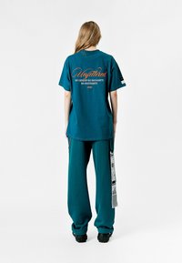 Camisa de gran tamaño en color verde azulado con texto gráfico en la parte trasera, combinada con pantalones anchos a juego que presentan una etiqueta lateral en contraste.