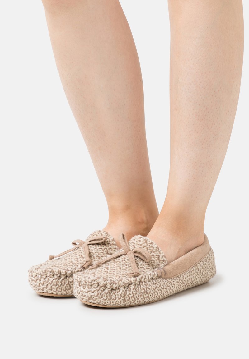 Gehaakte beige pantoffels met een zachte crèmekleurige voering, ronde neus en strikdetail aan de voorkant. Gelaagd geweven stof en platte zool.