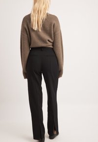 Hnedý pletený sveter s prenikavo širokými rukávmi, skombinovaný s čiernymi nohavicami s vysokým pásom, ktoré majú rozparky a zadné vrecká.