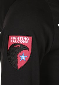 Musta paita, jossa on punainen kilpamerkki, jossa on musta kotka ja teksti "FIGHTING FALCONS" sinisen tähden yläpuolella, jonka sisällä on "TG".