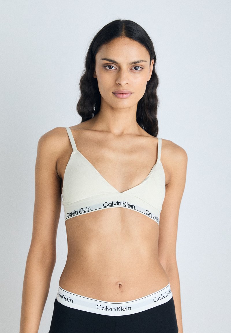 Svetlo bež braletka s tankimi naramnicami in globokim izrezom, ki ima črn pas z logotipom Calvin Klein na dnu. Gladka tekstura.