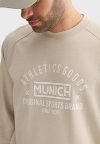Sweat-shirt beige avec un col rond et des manches raglan. Présente un texte imprimé blanc : "ATHLETICS GOODS MUNICH MARQUE DE SPORTS ORIGINALE DEPUIS 1939."