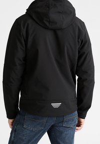 Veste softshell noire avec capuche, présentant une finition texturée, un design avec évent à l'arrière et un détail de bande réfléchissante au bas du dos.