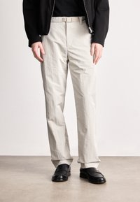 Απαλά γκρι, υφασμάτινα παν trousers με ίσια γραμμή και κρεμ ζώνη. Συνδυασμένα με μαύρα loafers, σε ουδέτερο φόντο.