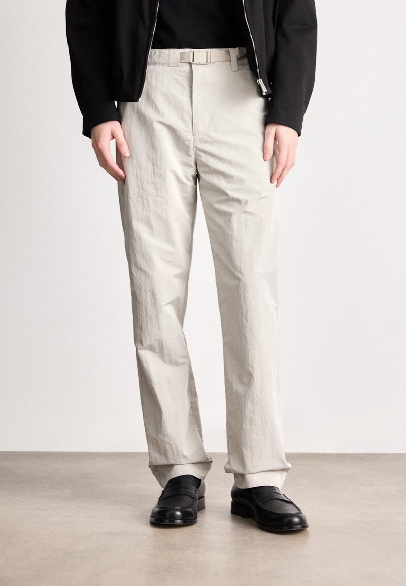 Απαλά γκρι, υφασμάτινα παν trousers με ίσια γραμμή και κρεμ ζώνη. Συνδυασμένα με μαύρα loafers, σε ουδέτερο φόντο.