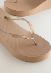 Paire de tongs beiges avec semelles texturées et brides en plastique transparent en forme de Y sur une surface claire.