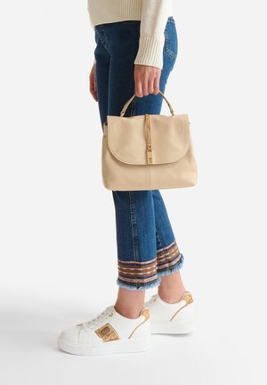 Persona con jeans corti blu con orli ricamati che tiene una borsa beige, indossando sneakers bianche con dettagli marroni.