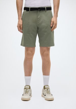 Πράσινα βαμβακερά σορτς με υφή, με μαύρη ζώνη. Συνδυάζονται με λευκές κάλτσες αστραγάλου και μπεζ αθλητικά παπούτσια. Απλή, ίσια γραμμή.