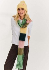 Kobieta o długich blond włosach, ubrana w żółtą dzianinową czapkę, białą koszulę z długim rękawem, kolorowy pasiassty szalik i ciemne spodnie na jednolitym tle.