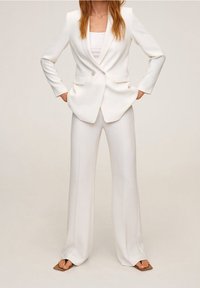 Žena v bílém přizpůsobeném sáčku s odpovídajícími širokými kalhotami a hnědými sandály s čtveratou špičkou, stojící s rukama v kapsách.