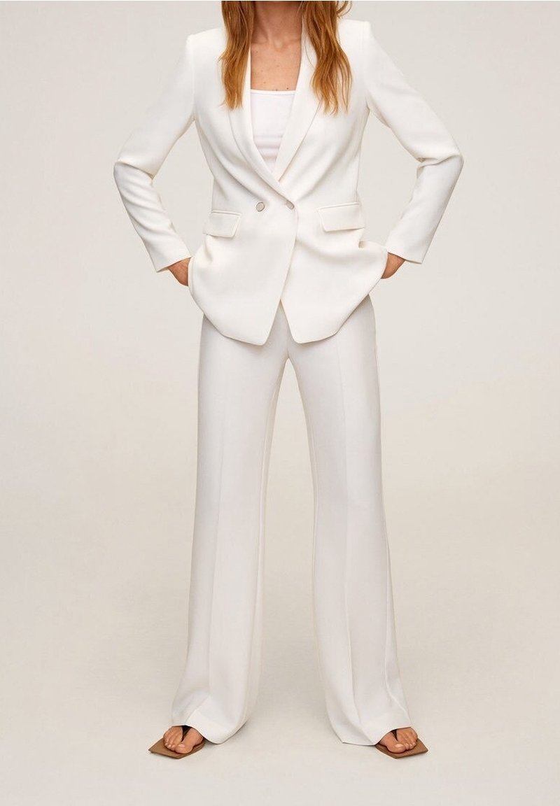 Žena v bílém přizpůsobeném sáčku s odpovídajícími širokými kalhotami a hnědými sandály s čtveratou špičkou, stojící s rukama v kapsách.