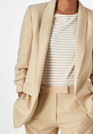 Beżowa lniana marynarka i pasujące do niej spodnie noszone na białej i beżowej koszuli w paski, ręce w kieszeniach, styl casual profesjonalny.