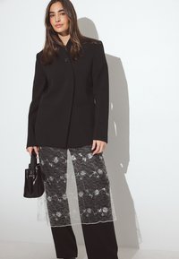 Zwarte op maat gemaakte blazer met een enkele knoop, gecombineerd met een sheer kanten rok met florale patronen over zwarte broeken. Houdt een zwarte handtas vast.