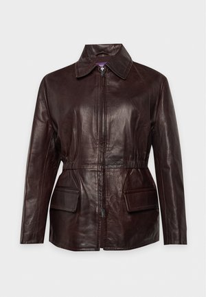 Une femme porte une veste en cuir marron foncé et une écharpe, avec un pull gris, un pantalon blanc et des chaussures marron, sur un fond uni.