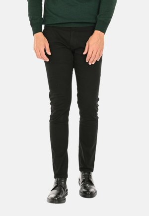 Pantaloni neri slim-fit realizzati in tessuto elasticizzato, con una superficie liscia e dettagli minimi, abbinati a scarpe in pelle nera lucida.