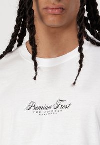 T-shirt bianco a maniche corte in cotone con logo stampato nero "Premium Frost, THE COLDEST WORLDWIDE" sul petto. Texture liscia.