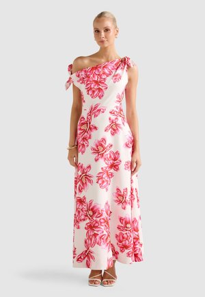 Donna che indossa un abito maxi bianco monospalla con grande stampa floreale rosa e dettagli legati sulla spalla, abbinato a sandali bianchi e gioielli dorati.