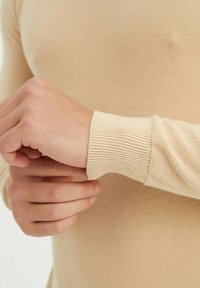 Chemise beige à manches longues en tricot avec des poignets côtelés, montrant un gros plan d'une main ajustant la manche. Motif texturé visible sur le tissu.