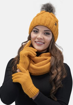 Mosterdgele gebreide beanie met pompon, bijpassende handschoenen en een dikke sjaal. De textuur is geribbeld en comfortabel, ontworpen voor warmte.