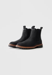 Botas Chelsea de cuero negro con suelas marrones, paneles laterales elásticos y tiradores, mostradas en un ligero ángulo sobre un fondo blanco.