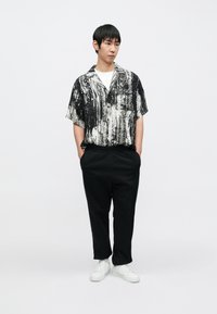 Schwarz-weiß gemustertes Kurzarmhemd mit abstraktem Muster, kombiniert mit schwarzen, zulaufenden Hosen und weißen Turnschuhen, vor neutralem Hintergrund präsentiert.