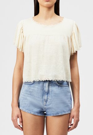 Frau trägt eine cremefarbene Kurzarmbluse mit Spitzenbesatz und hellblaue Jeansshorts, steht vor einem schlichten weißen Hintergrund.