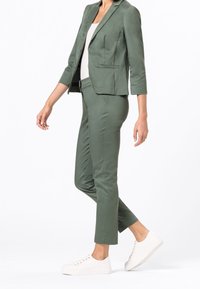 Zelený blazer a odpovídající zkrácené kalhoty z hladké látky, doplněné bílými teniskami. Má přiléhavý střih a minimalistický design.