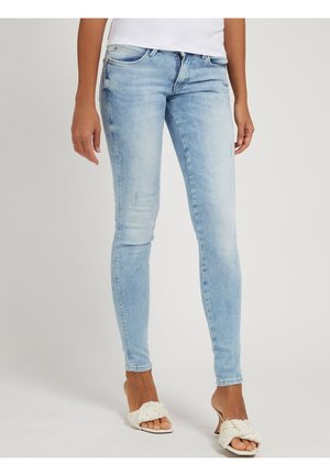 Femme portant un jean skinny bleu clair et des sandales blanches à talons avec des brides tressées, debout devant un fond uni.