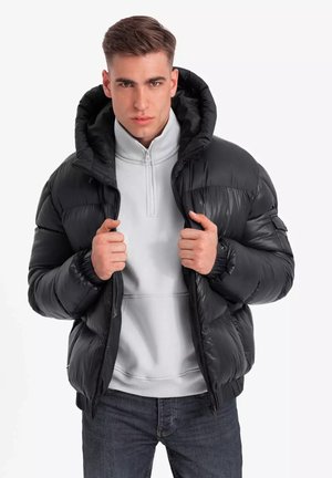 Giacca imbottita nera con un ampio cappuccio, chiusura con zip e polsini elastici. Indossata sopra un felpa grigia con zip e abbinata a jeans scuri.