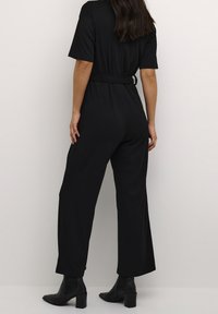 Mono negro de canalé con mangas cortas, cintura ceñida con cinturón y diseño de pierna ancha, combinado con botas ankle negras. Textura suave y estilo minimalista.