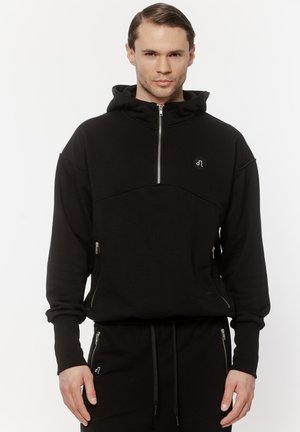 Mężczyzna w czarnej bluzie z kapturem i dopasowanych czarnych spodniach z detalami na zamkach, stojący na białym tle.