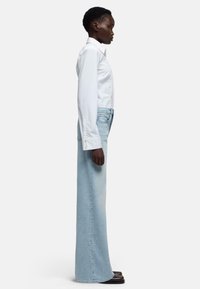 Camicia bianca a maniche lunghe abbinata a jeans a gamba larga di colore azzurro chiaro. I jeans hanno vita alta e un leggero scolorimento, completati da scarpe scure.