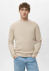 Beige geribbelde trui met een ronde hals en lange mouwen. Draagt een lichtblauwe spijkerbroek met een ontspannen pasvorm en subtiele textuur.