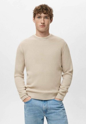 Beige geribbelde trui met een ronde hals en lange mouwen. Draagt een lichtblauwe spijkerbroek met een ontspannen pasvorm en subtiele textuur.