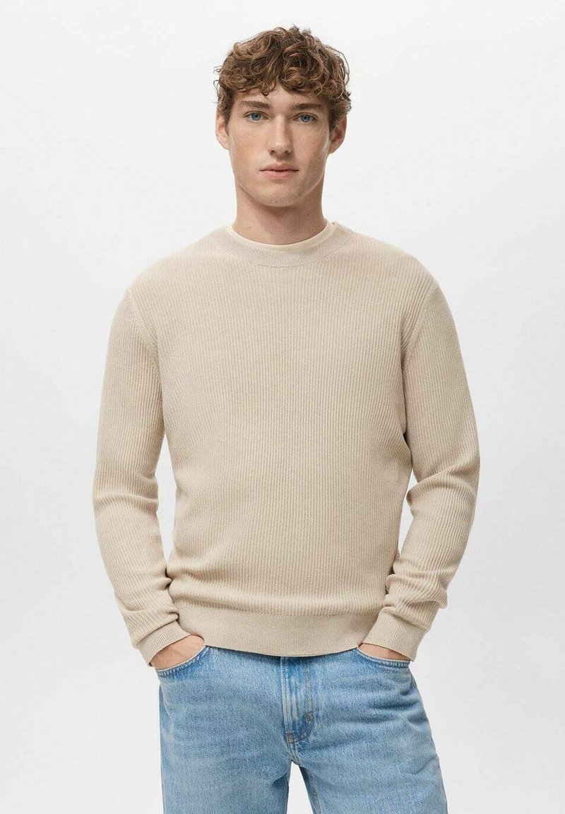 Beige geribbelde trui met een ronde hals en lange mouwen. Draagt een lichtblauwe spijkerbroek met een ontspannen pasvorm en subtiele textuur.