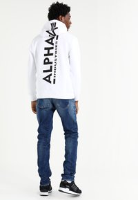 Witte hoodie met grote zwarte "ALPHA INDUSTRIES" tekst op de achterkant, gecombineerd met blauwe spijkerbroek en zwarte sneakers.