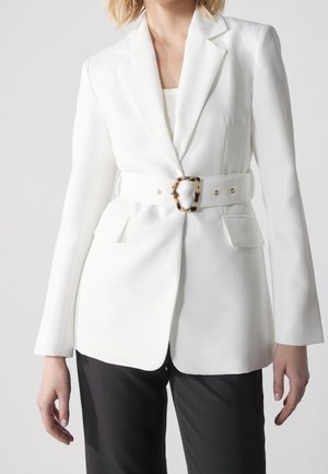 Vrouw draagt een op maat gemaakte witte blazer met brede riem met een schildpadgesp, gecombineerd met zwarte broek, tegen een effen achtergrond.