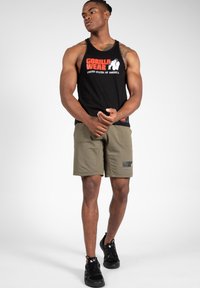 Črna majica brez rokavov z grafiko "GORILLA WEAR", v kombinaciji z olivno zelenimi kratkimi hlačami, ki imajo logotip. Model nosi črne športne �čevlje.
