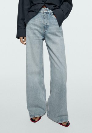 Frau trägt hellblaue Jeans mit weitem Bein, ein schwarzes Hemd mit Knöpfen, die Hände in den Taschen und offene schwarze und rote Sandalen.