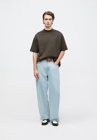 Ung mann som står iført en mørkebrun oversized kortermet skjorte, lyseblå vide jeans, brun belte og svart-hvite sko.