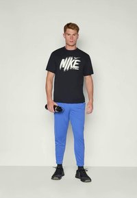 Zwarte Nike t-shirt met wit logo, blauwe sportbroek en zwarte sneakers. Het model houdt een waterfles vast en staat tegen een neutrale achtergrond.