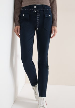 Persoon draagt donkerblauwe broek met klepzakken aan de voorkant en een knoopdetail, gecombineerd met witte sneakers en een bruin shirt met lange mouwen.