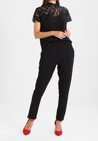 Frau trägt schwarzen Spitzenoberteil, schwarze, eng zulaufende Hose und rote spitze Absätze, steht vor weißem Hintergrund.