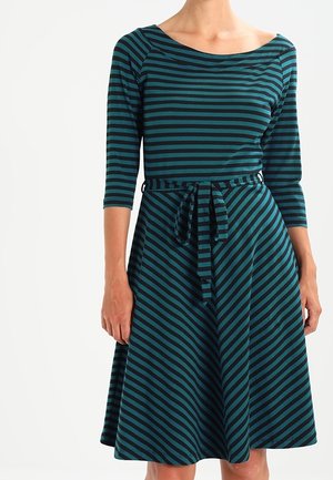 Kobieta w sukience w kolorach morskim i czarnym, z ukośnymi paskami, z rękawami do 3/4 długości i przewiązanym paskiem w talii, stojąca na białym tle.