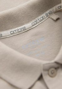 Beige bavlněná polo košile s rebrovaným límcem, obsahující štítek s n�ápisy "ombre" a "cotton line" v světle modrém písmu. Viditelný detail knoflíku.