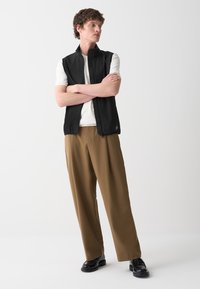 Gilet nero senza maniche con colletto alto, indossato sopra una camicia bianca, abbinato a pantaloni larghi marroni e scarpe nere. Design semplice.