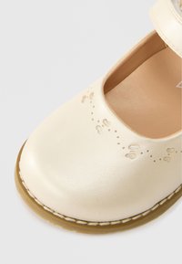 Kreemvärvi nahkjalats ümmarguse varbaga, lilledega perforatsioonidega kaunistatud ja ribaotsaga kinnitatud. Tekstuuriga tallaserv ja sile viimistlus.