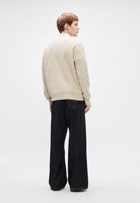 Pulover tricotat bej cu tiv ondulat, asortat cu blugi cu picioare largi de culoare albastru închis, care au buzunare la spate și detalii de cusătură.